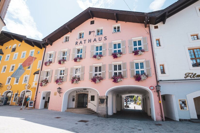 Rathaus Kitzbühel Ein farbenfrohes Rathaus in einer malerischen Straße. Die Fassade ist rosa mit Blumenfensterbänken und umgeben von weiteren schönen Gebäuden.