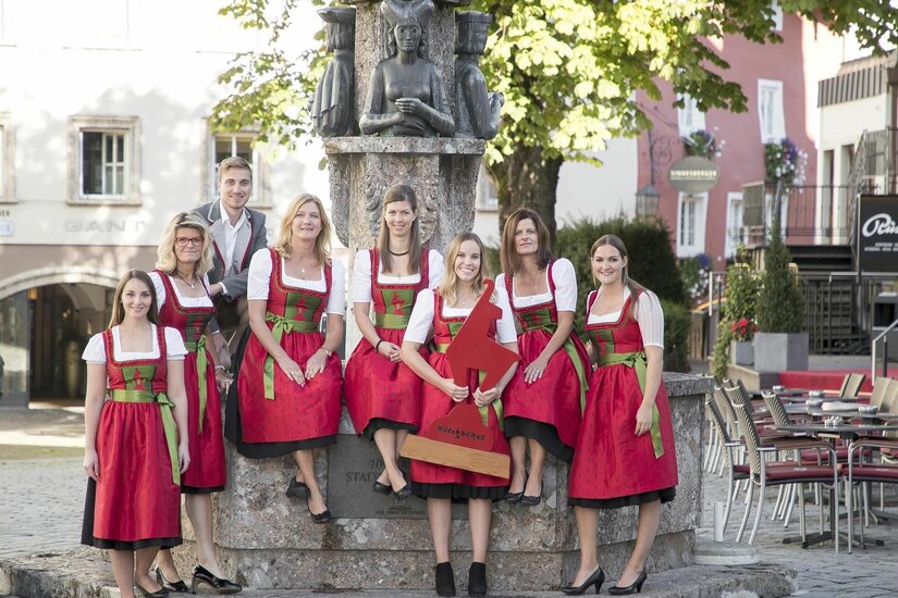 Kitzbühel Tourismus Team Eine Gruppe von Frauen in rot-grünen Dirndln und ein Mann posieren vor einem steinernen Brunnen in Kitzbühel.