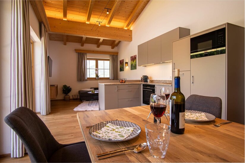 Apartment im Hof Zenzern Moderner Wohn-Essbereich mit Holzbalkendecke, Küchenzeile und gedecktem Holztisch mit Weingläsern.