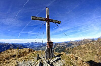 Ein hölzernes Gipfelkreuz auf einem grasbewachsenen Berggipfel vor blauem Himmel.