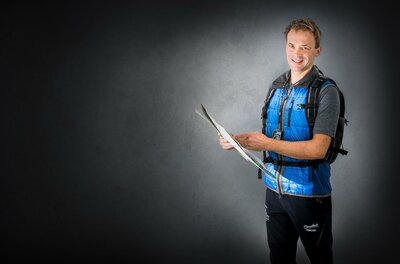 Ein junger Mann steht mit einem Rucksack und hält eine Landkarte in der Hand. Er lächelt und trägt eine blaue Weste.