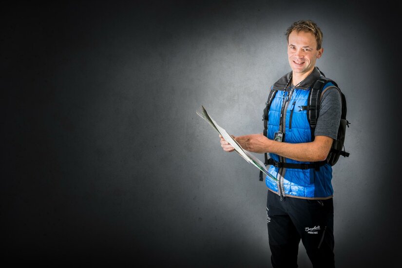 Reini Percht Ein junger Mann steht mit einem Rucksack und hält eine Landkarte in der Hand. Er lächelt und trägt eine blaue Weste.