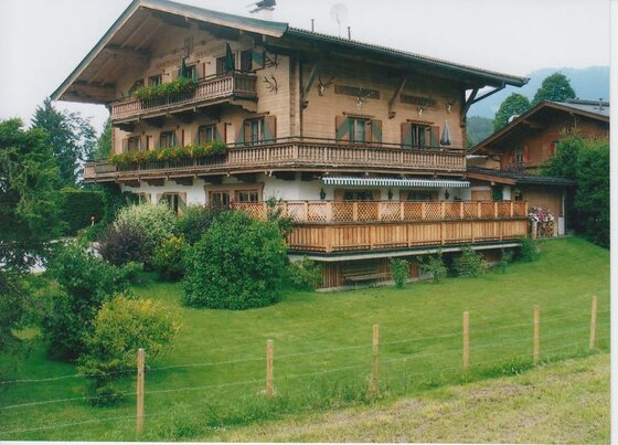 Hausfront Auracherhof Mehrstöckiges Holzhaus im alpinen Stil mit Balkonen, Blumenkästen und einer großen Terrasse vor einer Wiese.