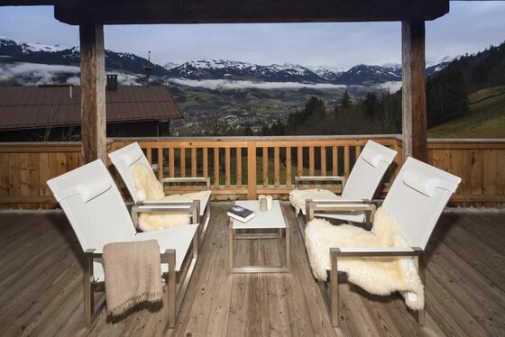 Holzbalkon mit vier weißen Sesseln und Blick auf schneebedeckte Berge und ein Tal.