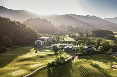 Grand Tirolia im Sommer Luftaufnahme eines Hotels mit Golfplatz vor bewaldeten Hügeln bei Gegenlicht.