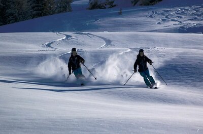 Zwei Skifahrer fahren nebeneinander durch tiefen Pulverschnee auf einem Hang.