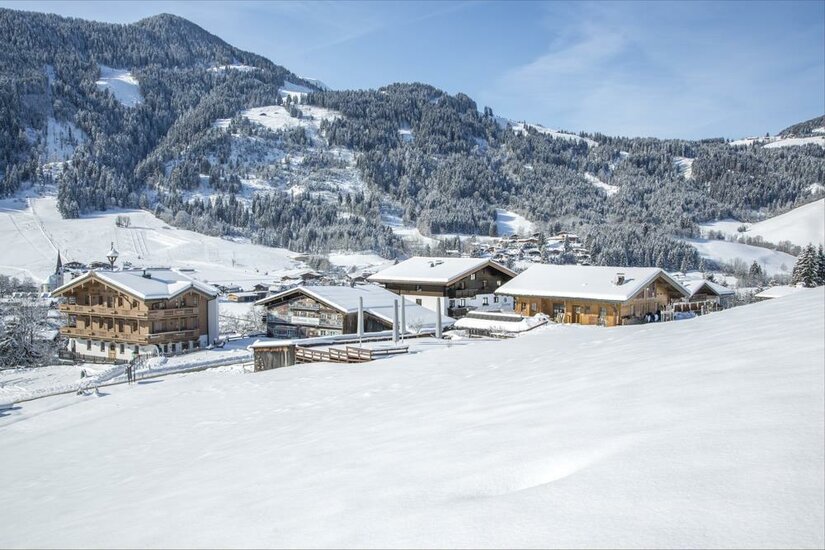 Verschneite Holzhäuser in einer alpinen Winterlandschaft vor bewaldeten Bergen.