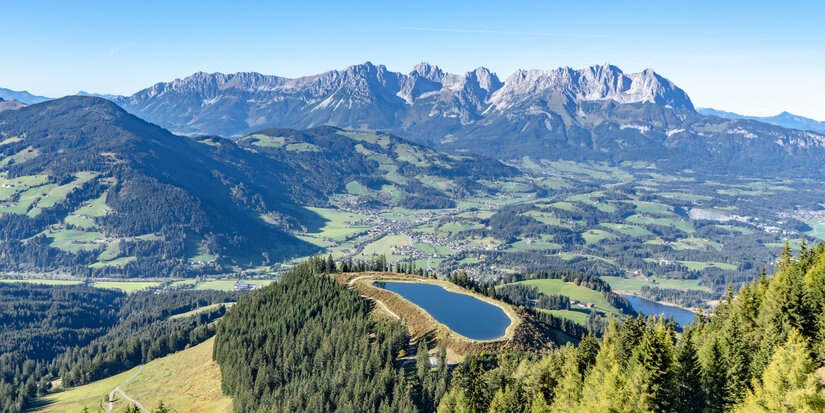 Ein atemberaubender Blick auf die Berge und Täler, mit einem klaren See auf einem bewaldeten Hügel im Vordergrund.