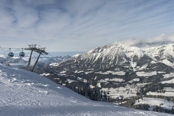 Eine schneebedeckte Berglandschaft mit einem Skilift und klaren, blauen Himmel. Im Hintergrund sind majestätische Berge und Täler zu sehen.