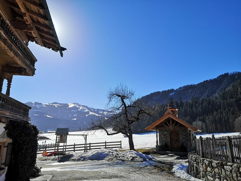 Alpines Holzhaus und kleine Kapelle vor verschneiter Berglandschaft unter blauem Himmel.