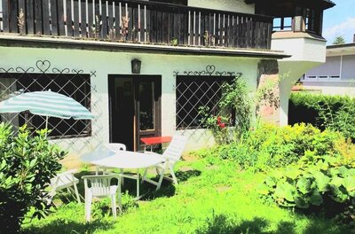 Garten mit weißen Kunststoffmöbeln, Sonnenschirm und einer Terrasse vor einem Haus mit Holzbalkon.