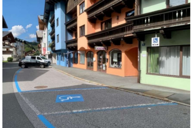Behindertenparkplatz - Im Grieß 29 Eine ruhige Straße mit bunten Gebäuden und einem klaren Himmel. Parkplätze sind deutlich markiert, einschließlich eines Behindertenparkplatzes.