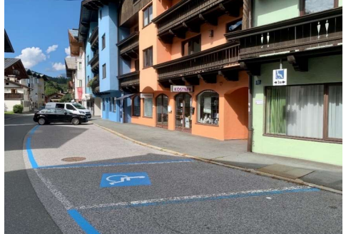 Behindertenparkplatz - Im Grieß 29 Eine ruhige Straße mit bunten Gebäuden und einem klaren Himmel. Parkplätze sind deutlich markiert, einschließlich eines Behindertenparkplatzes.