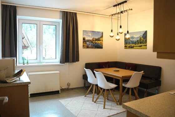 Eine gemütliche Essküche mit einem Holzschreibtisch und weißen Stühlen. An der Wand hängen zwei Bilder von Berglandschaften und es gibt große Fenster mit Vorhängen.
