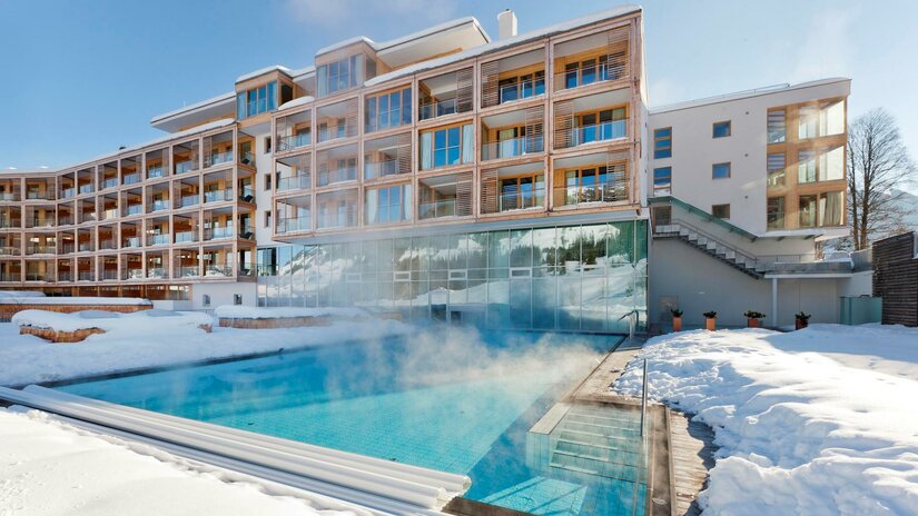 Winter-Außenpool Kempinski Hotel Dampfender Außenpool vor einem modernen Hotelgebäude in verschneiter Landschaft.