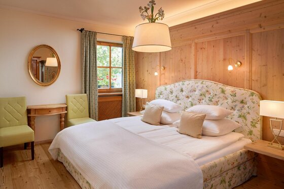 Schlafzimmer im Landsitz Römerhof Doppelbett mit geblümtem Kopfteil in einem Zimmer mit Holzvertäfelung, Spiegel und zwei grünen Stühlen.