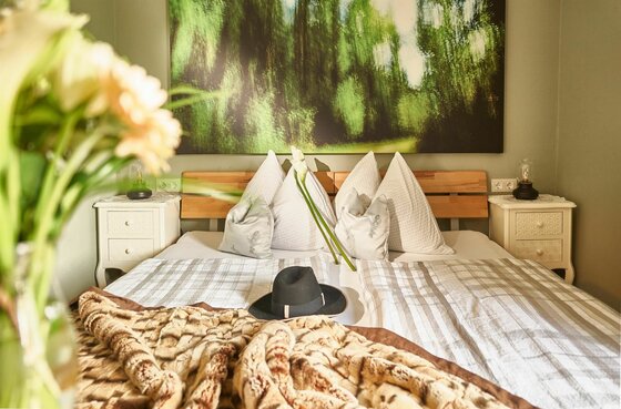 Doppelbett mit hellen Kissen, Hut auf der Decke und großem Waldbild an der Wand.
