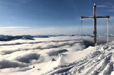 Gipfelkreuz auf dem verschneiten Hochetzkogel über einer geschlossenen Wolkendecke unter blauem Himmel.