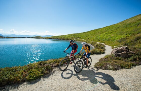 Mountainbiken am Bergsee Zwei Mountainbiker fahren auf einem Schotterweg an einem blauen Bergsee unter klarem Himmel.