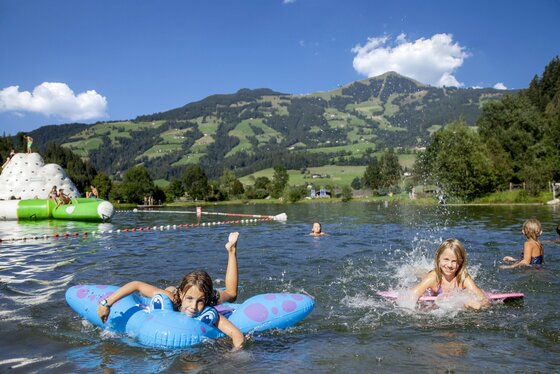 Salvena Badesee @Region Hohe Salve-Thomas Trinkl Kinder schwimmen in einem See mit bunten Schwimmringen. Im Hintergrund sieht man grüne Hügel und blauen Himmel.
