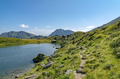 Ein schmaler Wanderpfad führt an einem Bergsee vorbei durch eine grüne Alpenlandschaft mit Berggipfeln im Hintergrund.