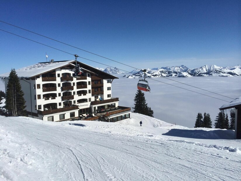 Berghotel Ehrenbachhöhe im Winter Ein mehrstöckiges Berghotel neben einer Skipiste und einem Sessellift über einer Wolkendecke im Winter.