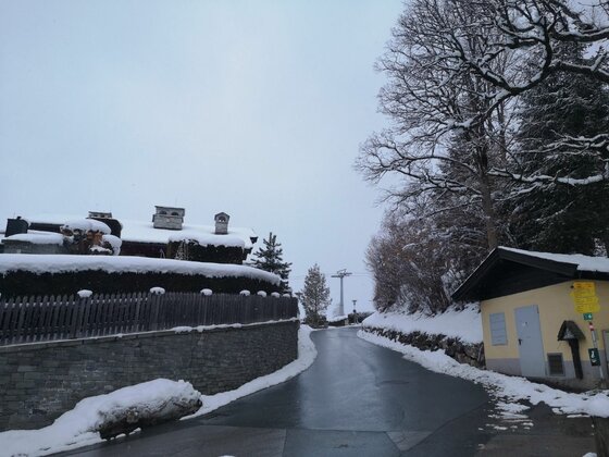 Bauernhaus-Museumsweg im Winter Verschneite Straße am Bauernhaus-Museumsweg mit Holzhäusern und Seilbahnmast im Hintergrund.