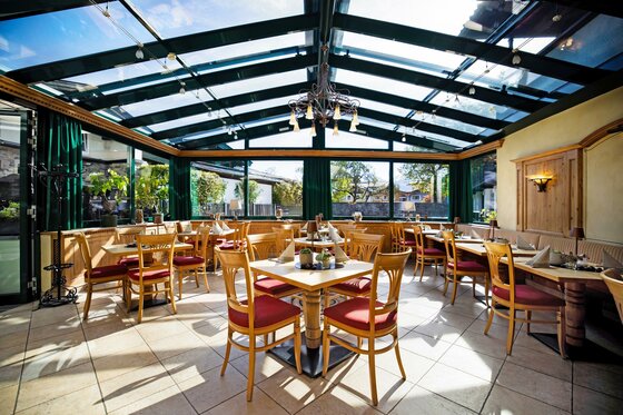 01Eggerwirt026 Ein helles Restaurant mit großen Fenstern und einem Glasdach. Die Tische sind sauber gedeckt und es gibt Holzstühle mit roten Polstern.