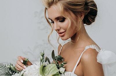 Galery Eine Braut mit elegantem Haar und einem wunderschönen Blumenstrauß steht vor einem neutralen Hintergrund. Ihr Kleid hat zarte Details und strahlt eine romantische Atmosphäre aus.