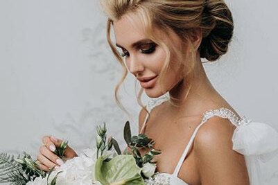 Brautstyling Hofmann Experience Braut mit hochgesteckten blonden Haaren und einem weißen Blumenstrauß vor hellem Hintergrund.