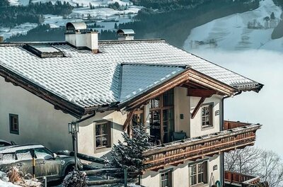 Residenz am Berg - Winter Alpines Wohnhaus mit Holzbalkon und schneebedecktem Dach vor verschneiter Bergkulisse.