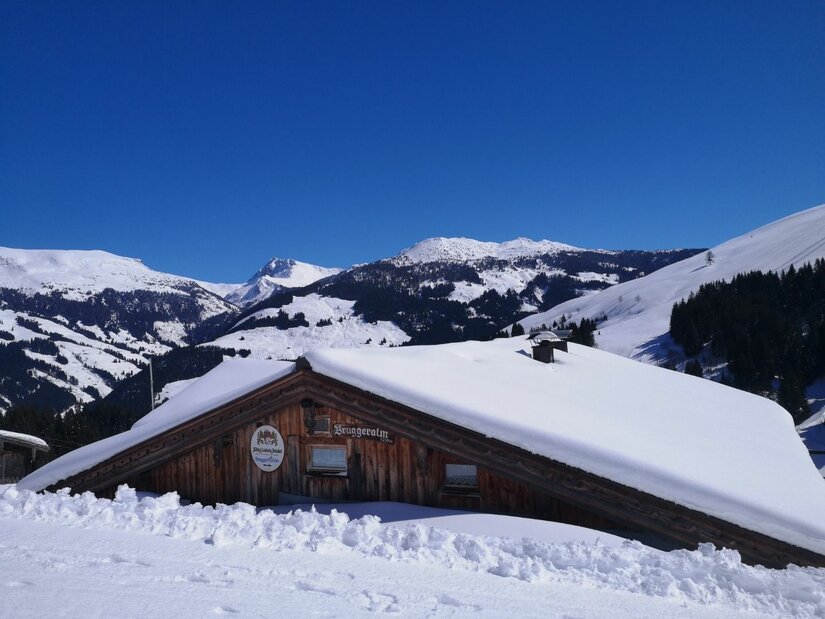 Bruggeralm im Winter Schneebedeckte Holzhütte vor verschneiten Berggipfeln unter blauem Himmel.