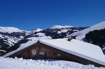 Schneebedeckte Holzhütte vor verschneiten Berggipfeln unter blauem Himmel.