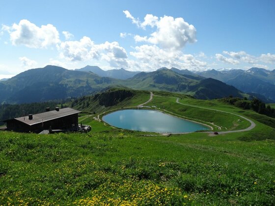 Raintal-Almen mit Bergsee Blick auf eine Almhütte und einen kleinen Bergsee inmitten grüner Almwiesen und Gebirgsketten.