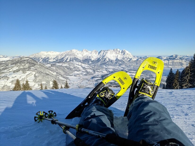 Person mit gelben Schneeschuhen sitzt im Schnee vor einem verschneiten Bergpanorama bei blauem Himmel.
