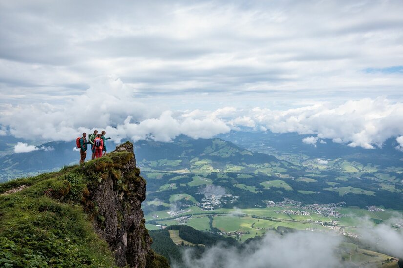 Vier Wanderer stehen auf einem grasbewachsenen Felsvorsprung mit Blick auf ein weites Tal unter Wolken.