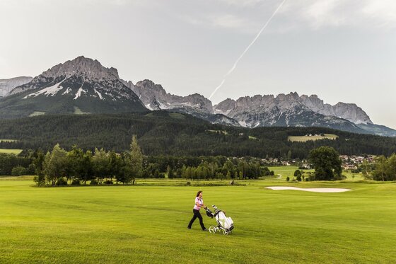 Eine Person geht mit einem Golfwagen über ein grünes Golfplatz. Im Hintergrund sind beeindruckende Berge zu sehen.