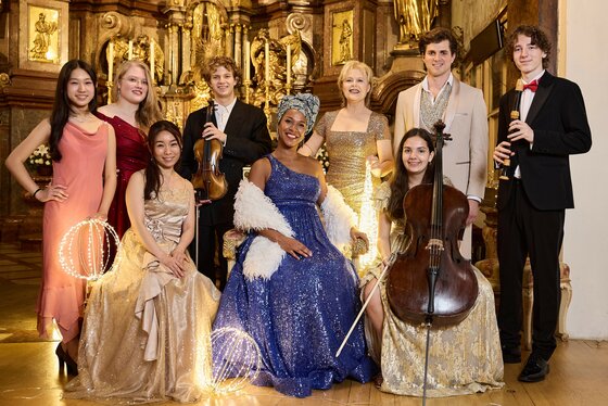 Acht Musiker in festlicher Kleidung mit Instrumenten vor einem goldenen Barockaltar.