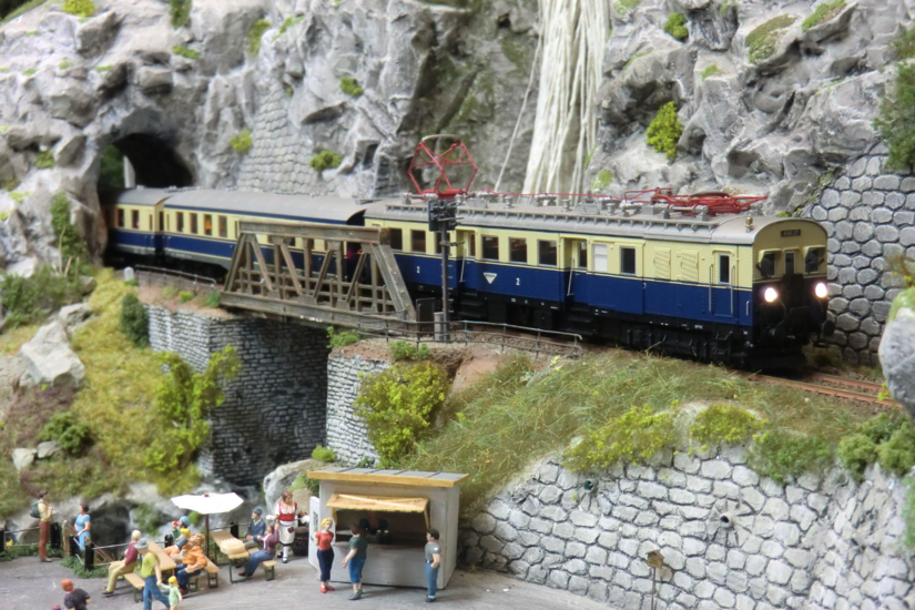 Modelleisenbahn mit blau-beigem Triebwagen auf einer Brücke vor einem Felstunnel und Miniaturfiguren.