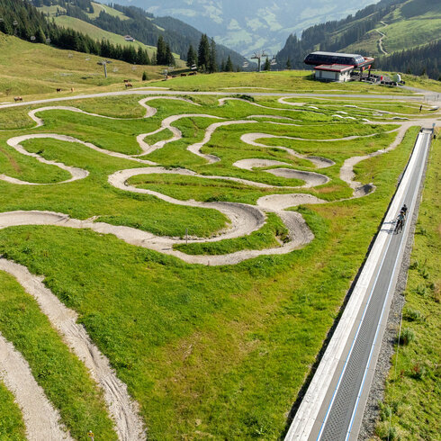 Eine kurvenreiche Strecke aus Schotter und Gras im MTB Family Area bei den 360 Flow Trails in Kitzbühel führt über eine grüne Wiese, umgeben von sanften Hügeln und Bäumen.
