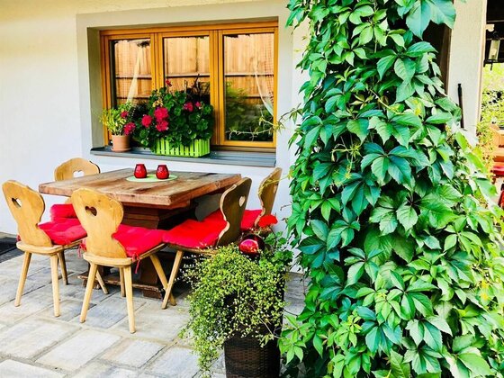 Terrasse Gästehaus Jochberg Holztisch mit vier Stühlen und roten Sitzkissen auf einer gepflasterten Terrasse neben einer begrünten Säule.