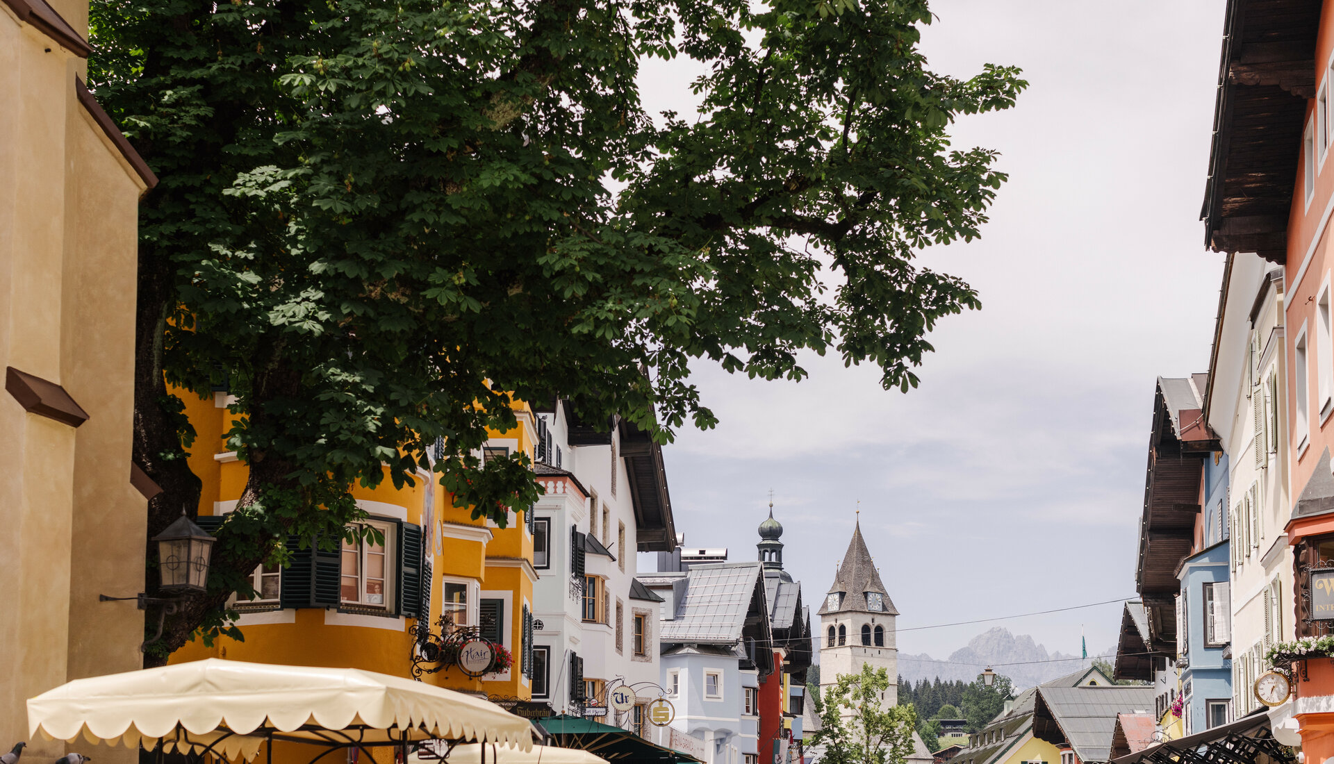 Bunte Häuserfassaden säumen die Straße in Kitzbühel, während ein großer Baum Schatten spendet und die Atmosphäre belebt.