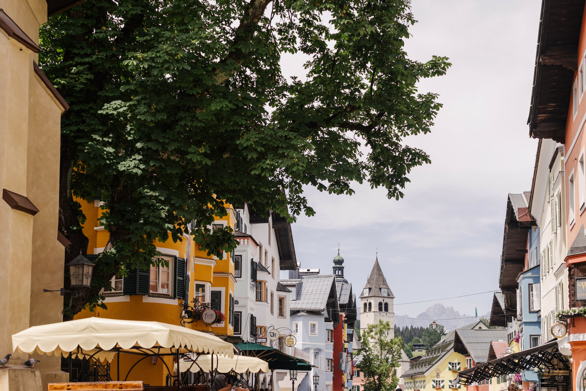 Bunte Häuserfassaden säumen die Straße in Kitzbühel, während ein großer Baum Schatten spendet und die Atmosphäre belebt.