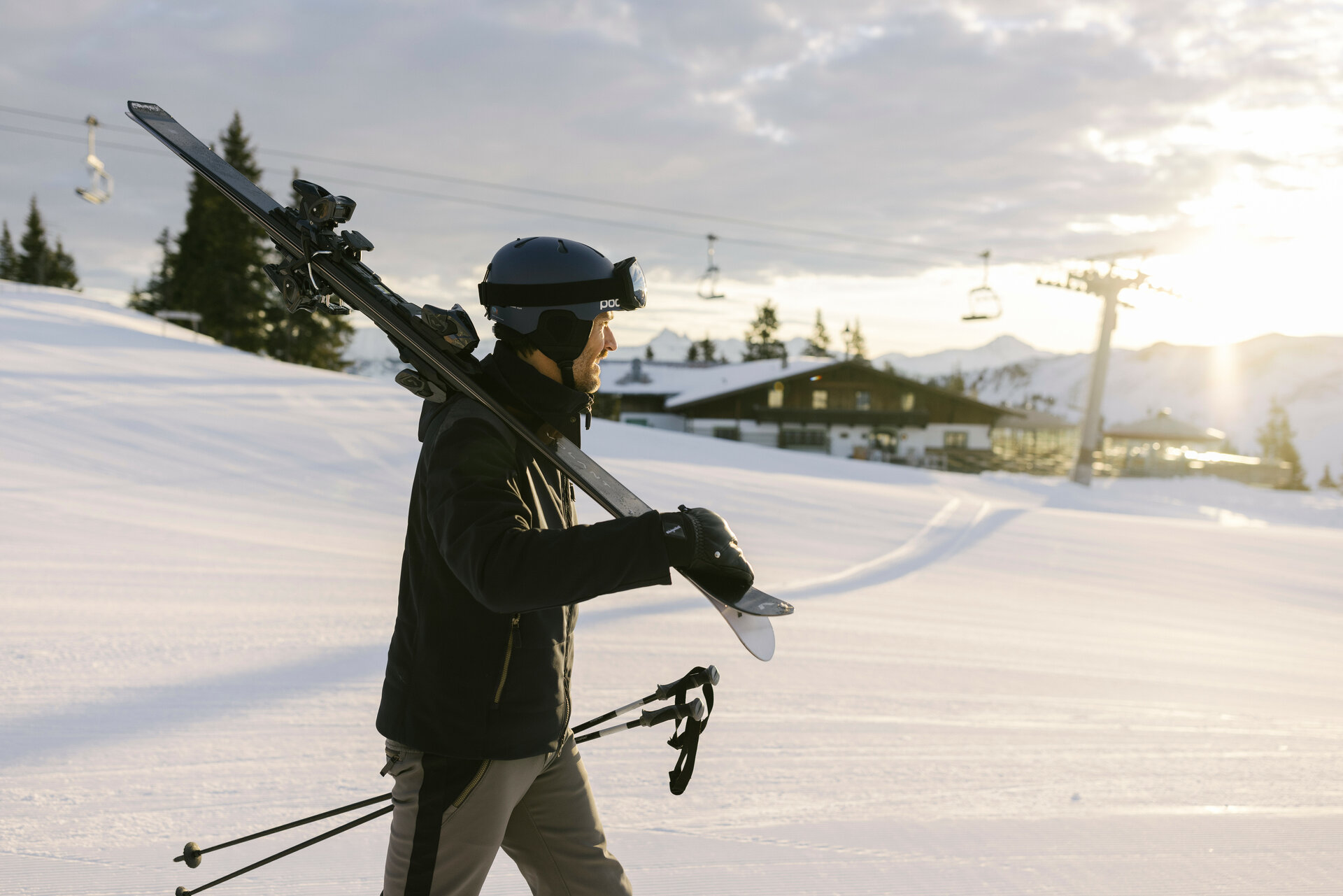 Der Mann mit Skiern auf der Schulter geht in Richtung einer Berghütte, umgeben von schneebedeckten Landschaften und Bäumen.