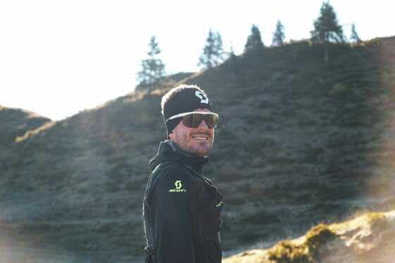 Trailrunner Paul Doyle Ein Mann mit Sportbrille, Stirnband und schwarzer Sportjacke vor einem bewaldeten Berghang.