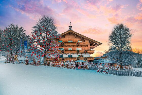 Ein schneebedecktes Haus in den Bergen, umgeben von winterlichen Bäumen. Der Himmel leuchtet in sanften Farben bei Sonnenuntergang.