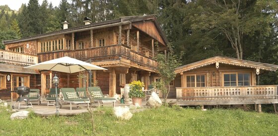 Außenansicht Berghütten Sonnschwendt Zweistöckige Holzhütte mit Terrasse, Sonnenliegen, Sonnenschirm und Grill vor einem Waldrand.