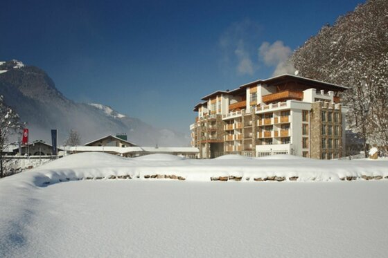 Mehrstöckiges Hotelgebäude mit Holzbalkonen in einer verschneiten Alpenlandschaft unter blauem Himmel.