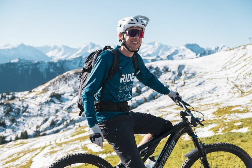 Mountainbiker in den Kitzbüheler Alpen Mountainbiker mit Helm und Rucksack auf einem Trail vor verschneiten Berggipfeln.
