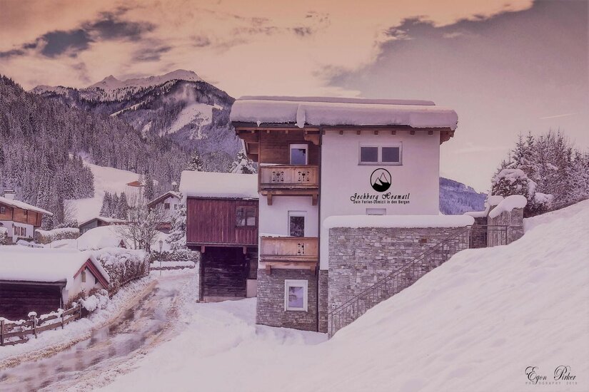 Hoamatl außen Schneebedecktes mehrstöckiges Ferienhaus mit Holzbalkonen vor bewaldeten Bergen im Winter.